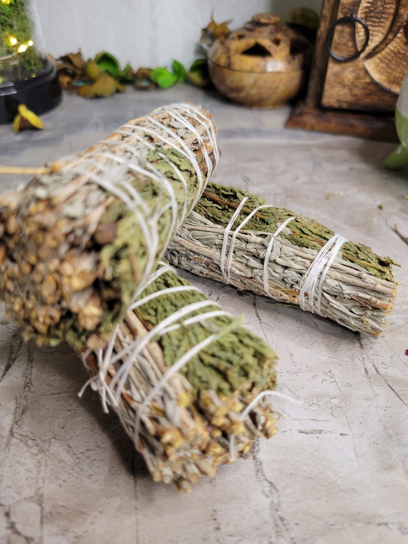 Pu&ograve; includere: Due mazzi di erbe secche legate con spago. Un mazzo &egrave; una miscela di salvia e cedro, l'altro &egrave; solo salvia. I mazzi sono su una superficie grigia.