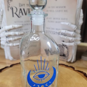 May include: A clear glass bottle with a spherical stopper featuring a blue graphic print of an eye centered within a crescent moon and radiating lines. The bottle has a rectangular body with rounded edges and a narrow neck, standing upright.
