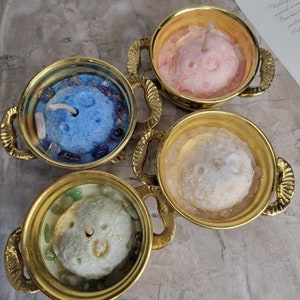 May include: Four decorative candles in gold-colored teacups. Each candle has a unique color: blue, pink, white, and green. The candles are surrounded by small stones and glitter. The teacups have ornate handles.
