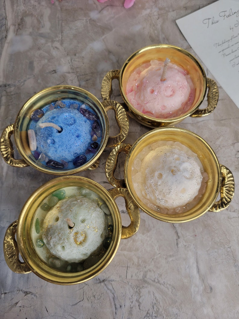 May include: Four decorative candles in gold-colored teacups. Each candle has a unique color: blue, pink, white, and green. The candles are surrounded by small stones and glitter. The teacups have ornate handles.
