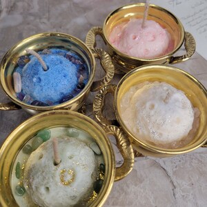 May include: Four decorative candles in gold-colored holders. Each candle is a different color: blue, pink, white, and green. The candles have a textured surface and are surrounded by small stones. The holders have ornate handles.
