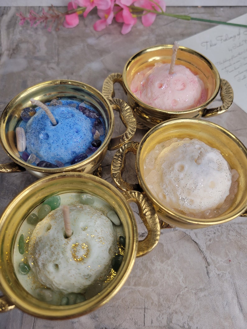 May include: Four decorative candles in gold-colored holders. Each candle is a different color: blue, pink, white, and green. The candles have a textured surface and are surrounded by small stones. The holders have ornate handles.