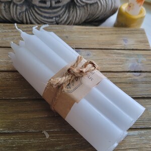May include: A bundle of six white taper candles secured with a brown paper band and tied with a natural jute twine bow. The paper band features a printed logo with a botanical leaf illustration and the partial text MYA P.