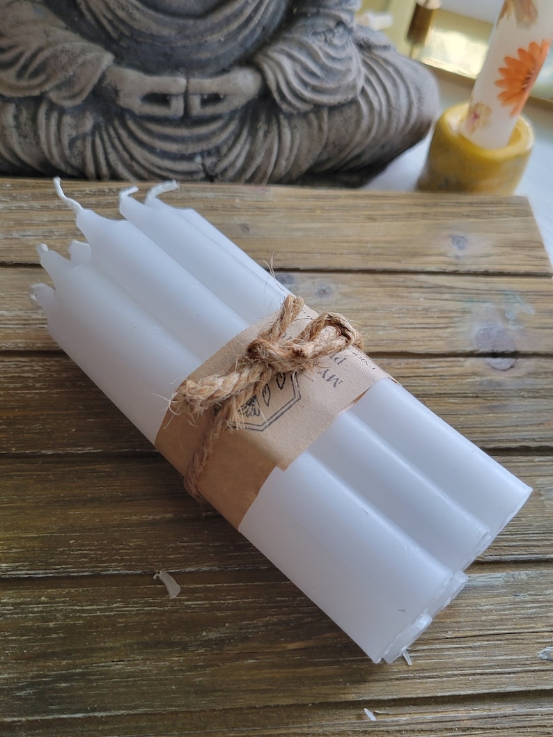 May include: A bundle of six white taper candles secured with a brown paper band and tied with a natural jute twine bow. The paper band features a printed logo with a botanical leaf illustration and the partial text MYA P.