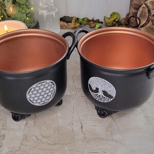 May include: Two black metal cauldrons with copper interiors and decorative white designs. One cauldron features a flower of life design, while the other has a tree of life design.