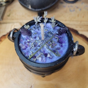 May include: A black cauldron-shaped candle filled with lavender-colored wax, amethyst crystals, and dried lavender buds. The candle has a central wick and is decorated with glitter. The cauldron has two metal handles and sits on a wooden surface.