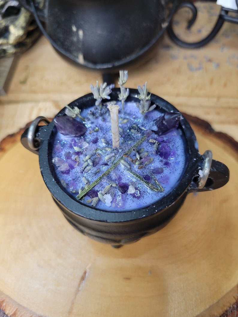 May include: A black cauldron-shaped candle filled with lavender-colored wax, amethyst crystals, and dried lavender buds. The candle has a central wick and is decorated with glitter. The cauldron has two metal handles and sits on a wooden surface.