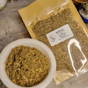 May include: A close-up of a small bag of dried Agrimony herb. The bag is labeled with the name of the herb, its magical properties, and its planetary association. The herb is also shown in a white bowl.