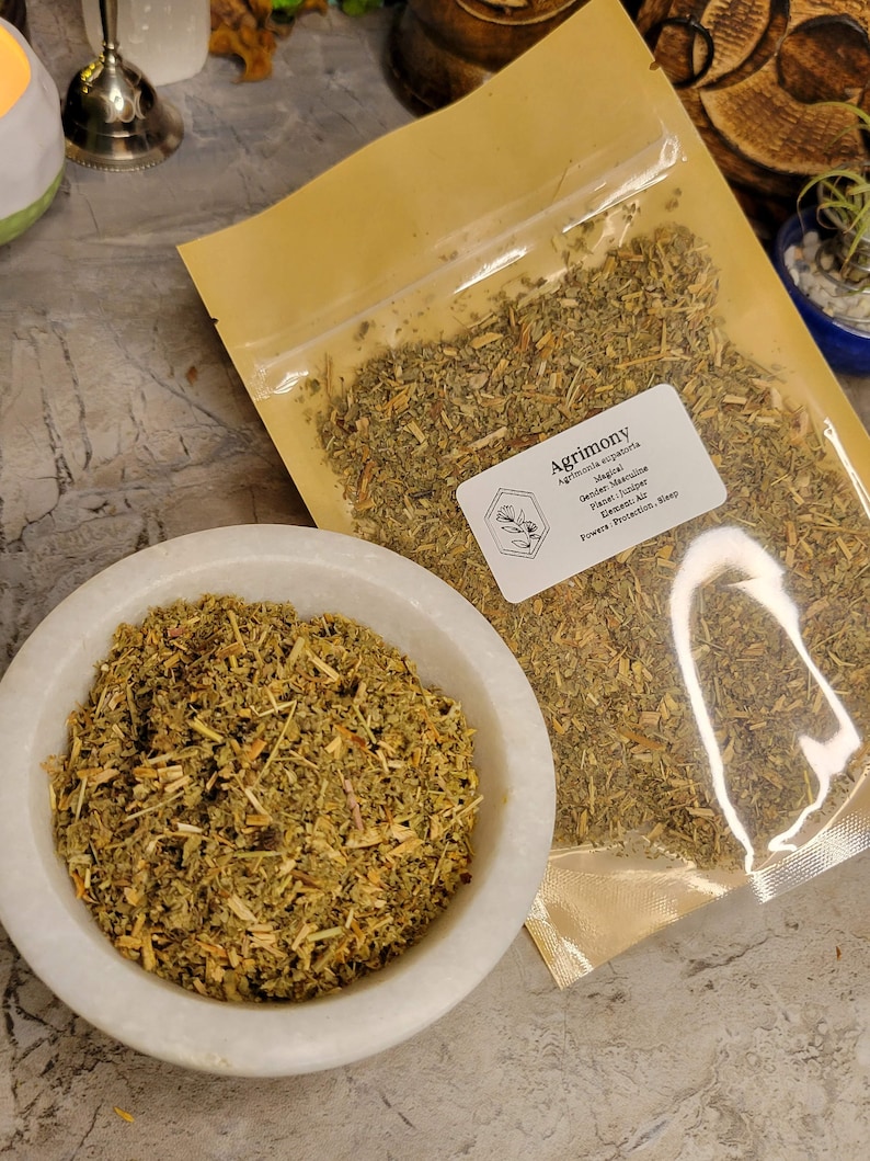 May include: A close-up of a small bag of dried Agrimony herb. The bag is labeled with the name of the herb, its magical properties, and its planetary association. The herb is also shown in a white bowl.