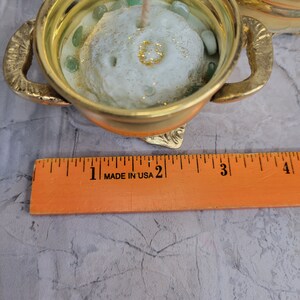 May include: A gold-colored candle holder with handles, filled with a light green candle. The candle has a textured surface and is decorated with green stones and gold glitter. A ruler that says "MADE IN USA" is in the foreground.