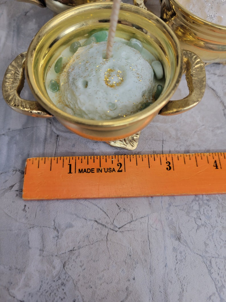 May include: A gold-colored candle holder with handles, filled with a light green candle. The candle has a textured surface and is decorated with green stones and gold glitter. A ruler that says "MADE IN USA" is in the foreground.