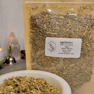 May include: A close-up of a bag of dried Agrimony herb. The bag is labeled with the name of the herb, its magical properties, and its gender, planet, element, and powers. The herb is a light green color and has a slightly brown tint.