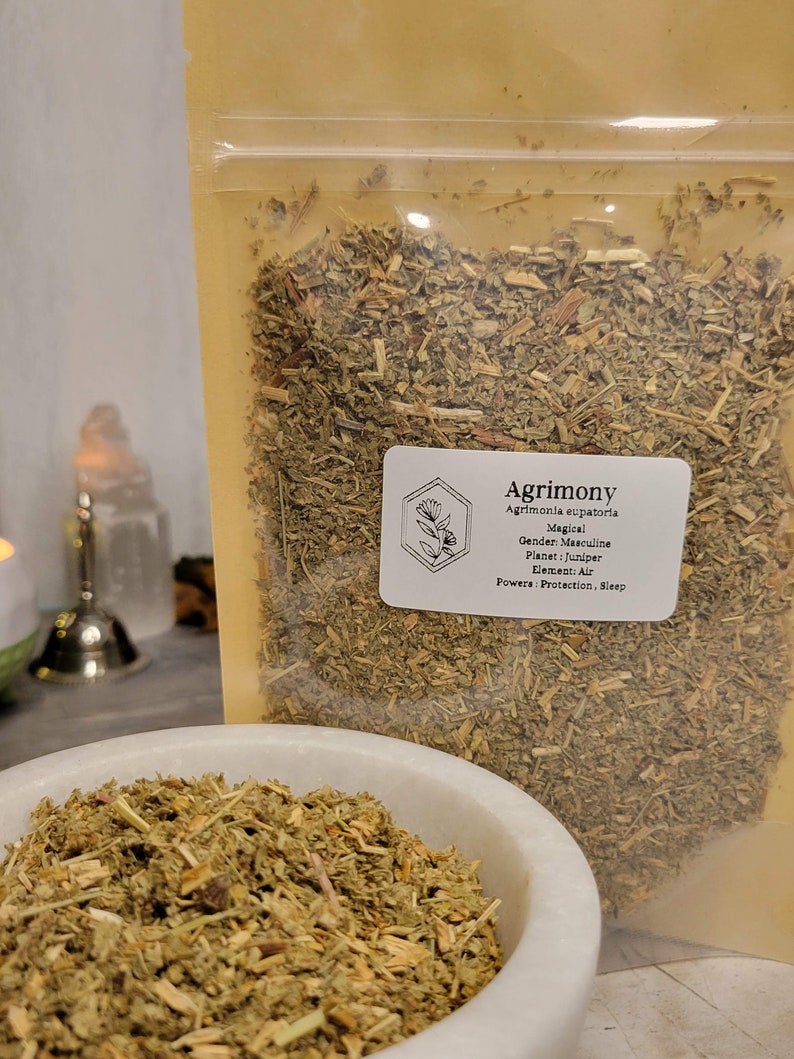 May include: A close-up of a bag of dried Agrimony herb. The bag is labeled with the name of the herb, its magical properties, and its gender, planet, element, and powers. The herb is a light green color and has a slightly brown tint.