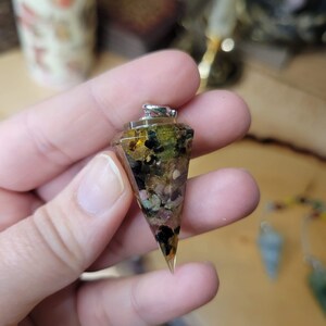 May include: A clear crystal pyramid pendant with a silver bail. The pyramid is filled with various small crystals, including green, brown, pink, and black.
