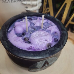 May include: A black cauldron-shaped candle with a lavender-colored wax interior. The wax features moon and star shapes, glitter, and crystals. Two wicks are visible. The cauldron sits on a wooden surface. The text "THEN THE BIRD SAID "NEVERMORE"" is visible in the background.