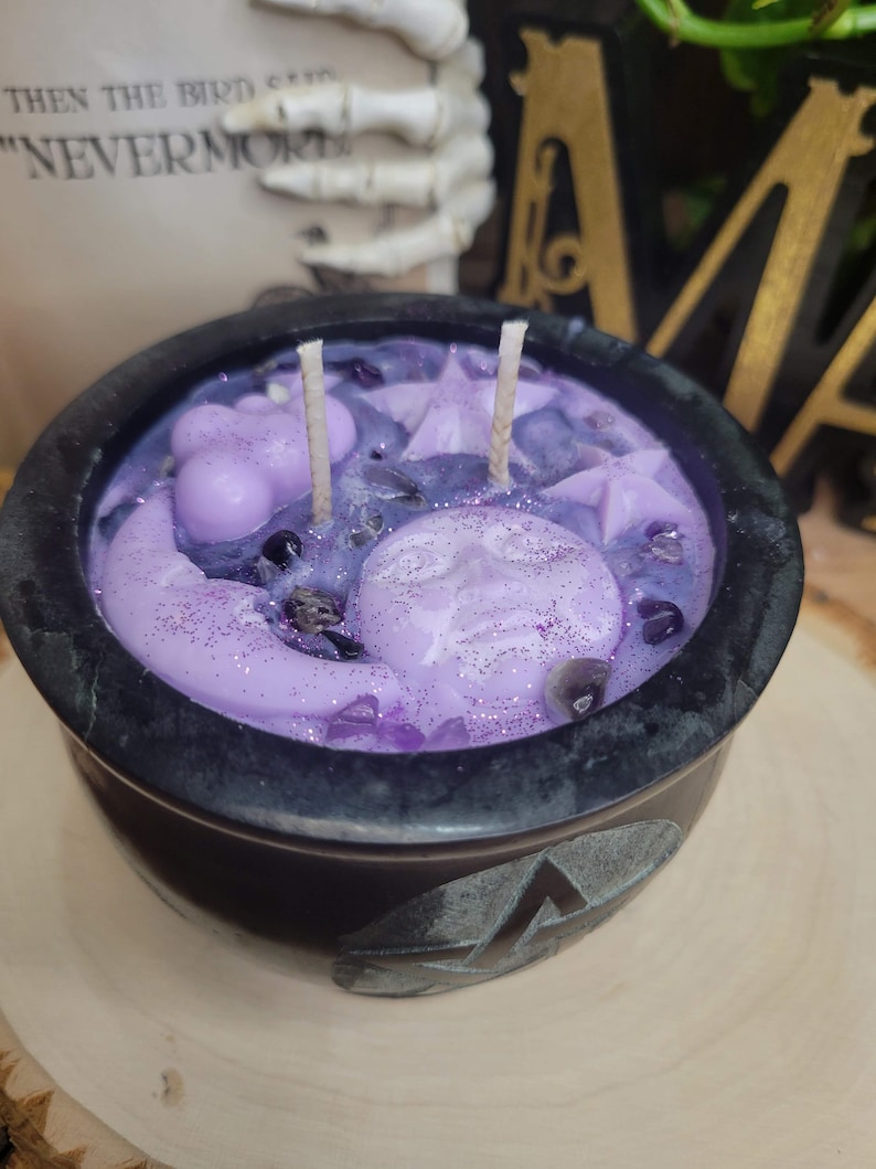May include: A black cauldron-shaped candle with a lavender-colored wax interior. The wax features moon and star shapes, glitter, and crystals. Two wicks are visible. The cauldron sits on a wooden surface. The text "THEN THE BIRD SAID "NEVERMORE"" is visible in the background.