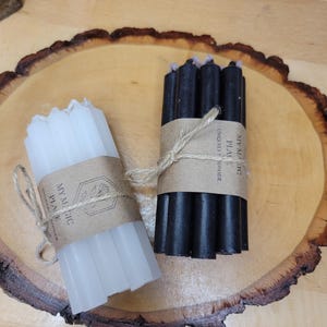 May include: Two bundles of thin, cylindrical candles, one white and one black, each wrapped in a brown paper band tied with twine. The paper bands feature the text My Magic Place and Uniquely Handmade. The candles are arranged in small groups.