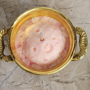 May include: A pink candle with glitter and crystals in a gold-colored bowl with handles. The candle has a light pink color with a glittery surface and is surrounded by clear crystals. The bowl has a decorative, textured handle.