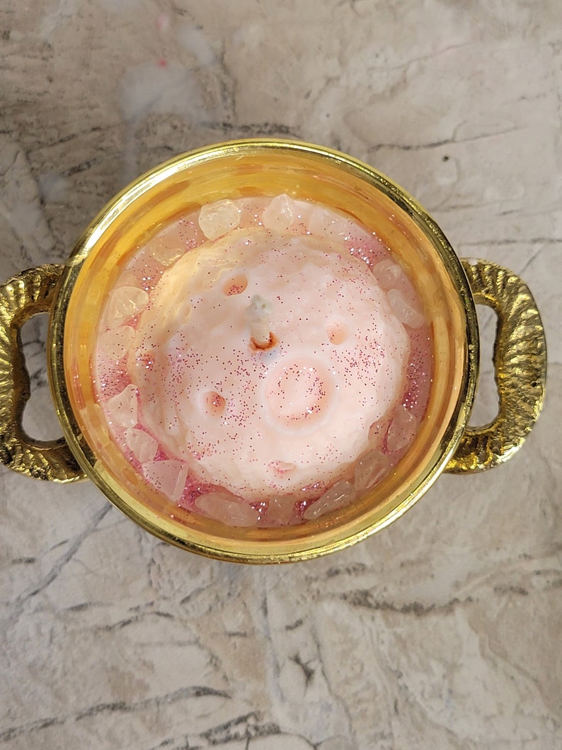 May include: A pink candle with glitter and crystals in a gold-colored bowl with handles. The candle has a light pink color with a glittery surface and is surrounded by clear crystals. The bowl has a decorative, textured handle.