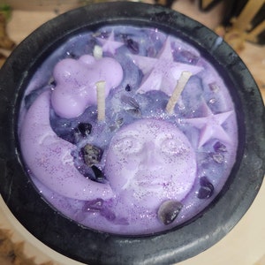 May include: A purple candle in a black cauldron-shaped container. The candle features a crescent moon with a face, stars, and a cloud. The candle is decorated with glitter and small, dark purple crystals.