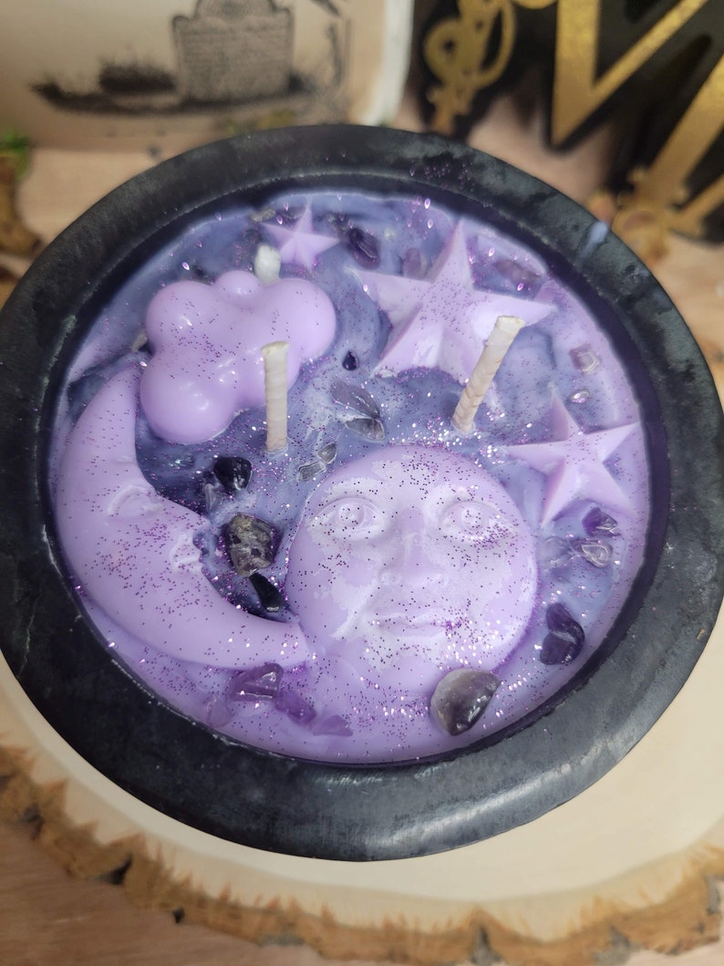 May include: A purple candle in a black cauldron-shaped container. The candle features a crescent moon with a face, stars, and a cloud. The candle is decorated with glitter and small, dark purple crystals.