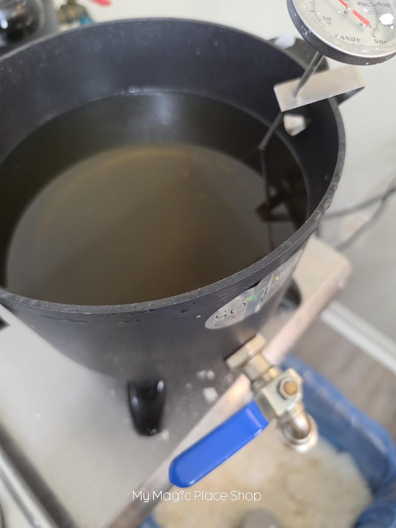May include: A black metal melting pot filled with liquid, featuring a silver candy thermometer clipped to the rim and a metal spigot with a blue handle attached to the side. The pot has a circular base and a smooth, dark exterior finish.