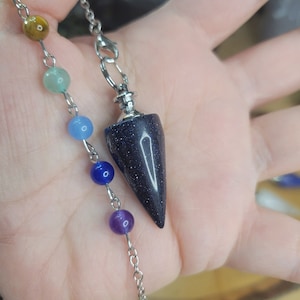 May include: A pendulum with a dark blue, speckled, cone-shaped stone. The pendulum is attached to a silver chain and is accompanied by a strand of colorful beads. The beads are in shades of yellow, brown, green, blue, and purple.