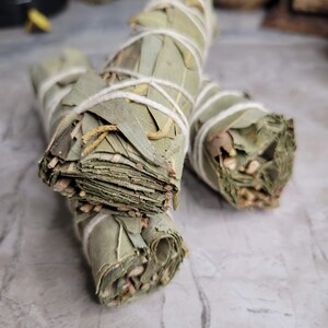 May include: Three bundles of dried eucalyptus leaves tied together with twine. The bundles are arranged on a light gray surface.