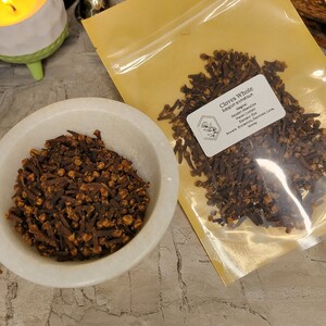 Puede incluir: Un primer plano de una bolsa de clavos enteros, una especia popular utilizada en la cocina y por sus propiedades medicinales. La bolsa est&aacute; etiquetada como "Clavos enteros" y enumera el nombre bot&aacute;nico "Syzygium aromaticum". La etiqueta tambi&eacute;n menciona los usos m&aacute;gicos de los clavos, incluida la protecci&oacute;n, el exorcismo, el amor y el dinero.