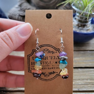 May include: A pair of silver dangle earrings with a rainbow of gemstones. The earrings are on a brown card with the text "Uniquely Handmade. Limited Edition."