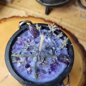 May include: A black cauldron-shaped candle with a lavender-colored wax, decorated with amethyst crystals, dried lavender, and a single wick. The candle has silver metal handles and is set on a wooden surface.