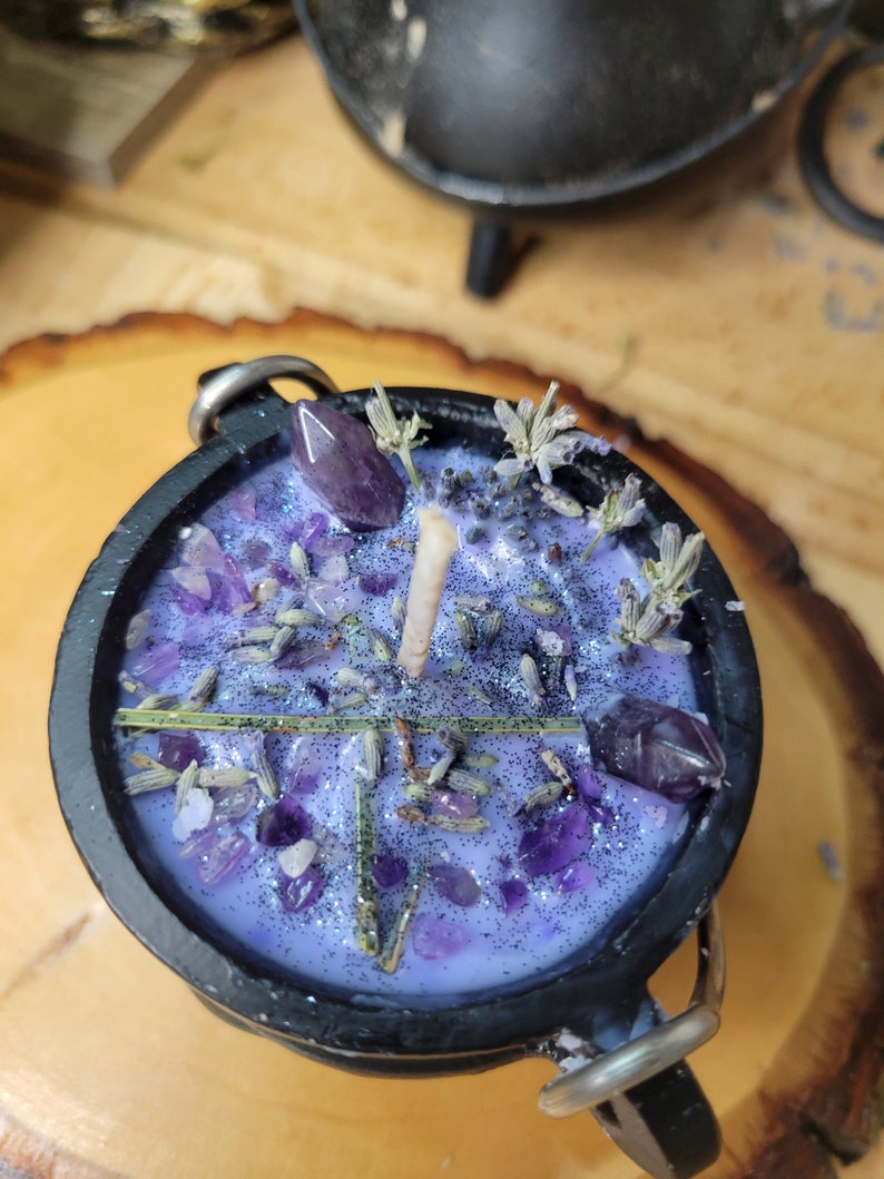 May include: A black cauldron-shaped candle with a lavender-colored wax, decorated with amethyst crystals, dried lavender, and a single wick. The candle has silver metal handles and is set on a wooden surface.