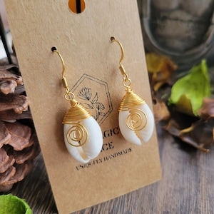 May include: A pair of handcrafted earrings featuring white cowrie shells wrapped in gold-colored wire. The earrings hang from gold-colored ear wires. The earrings are displayed on a brown card with the text "MAGIC PLACE UNIQELY HANDMADE".