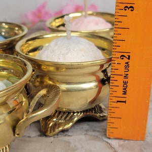 May include: Close-up of several small, gold-colored candle holders, each containing a small, round candle. The candles are various colors, including white and pink. The candle holders are ornate, with decorative bases. A ruler is in the frame, marked "MADE IN USA".