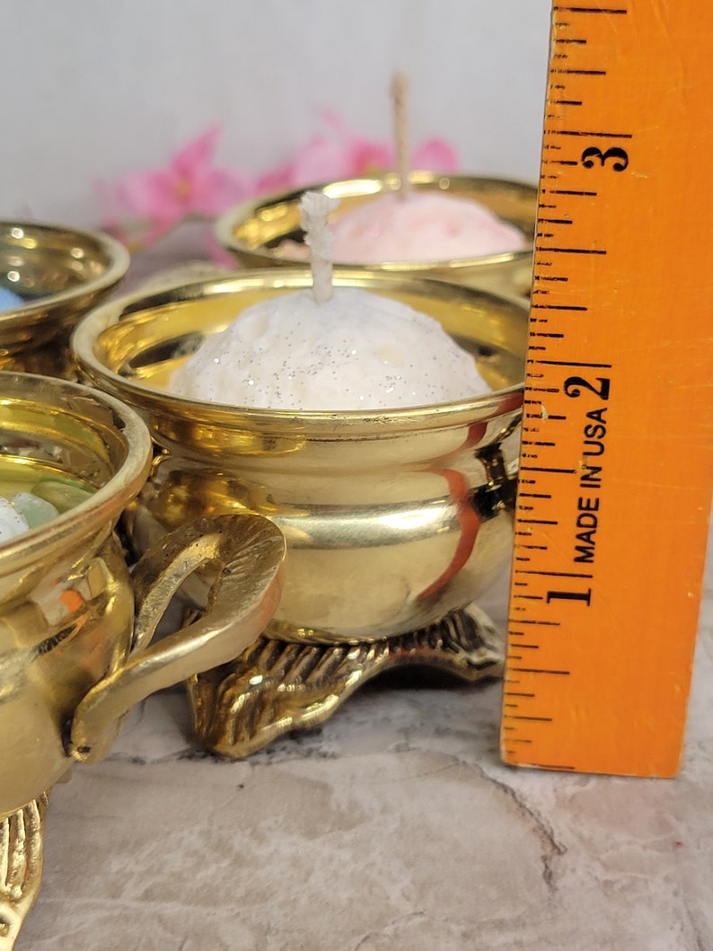 May include: Close-up of several small, gold-colored candle holders, each containing a small, round candle. The candles are various colors, including white and pink. The candle holders are ornate, with decorative bases. A ruler is in the frame, marked "MADE IN USA".