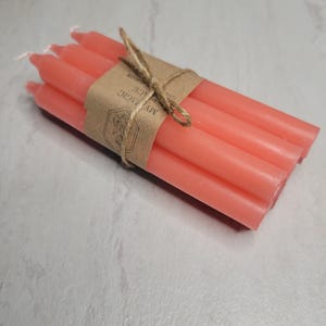 May include: A bundle of four coral-colored taper candles tied together with a brown paper band and twine. The paper band features a hexagonal logo with a floral illustration and the printed text MY MAGIC PLACE.