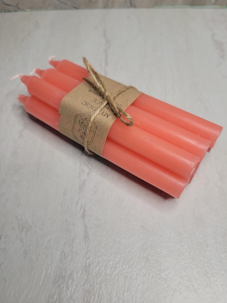 May include: A bundle of four coral-colored taper candles tied together with a brown paper band and twine. The paper band features a hexagonal logo with a floral illustration and the printed text MY MAGIC PLACE.