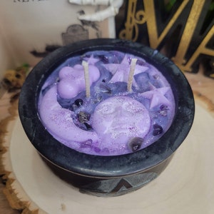May include: A black cauldron-shaped candle with a lavender-colored wax. The candle features a moon, stars, and cloud-shaped wax accents. The candle sits on a wooden surface. The text "THEN THE BIRD SAID "NEVERMORE" is visible in the background.