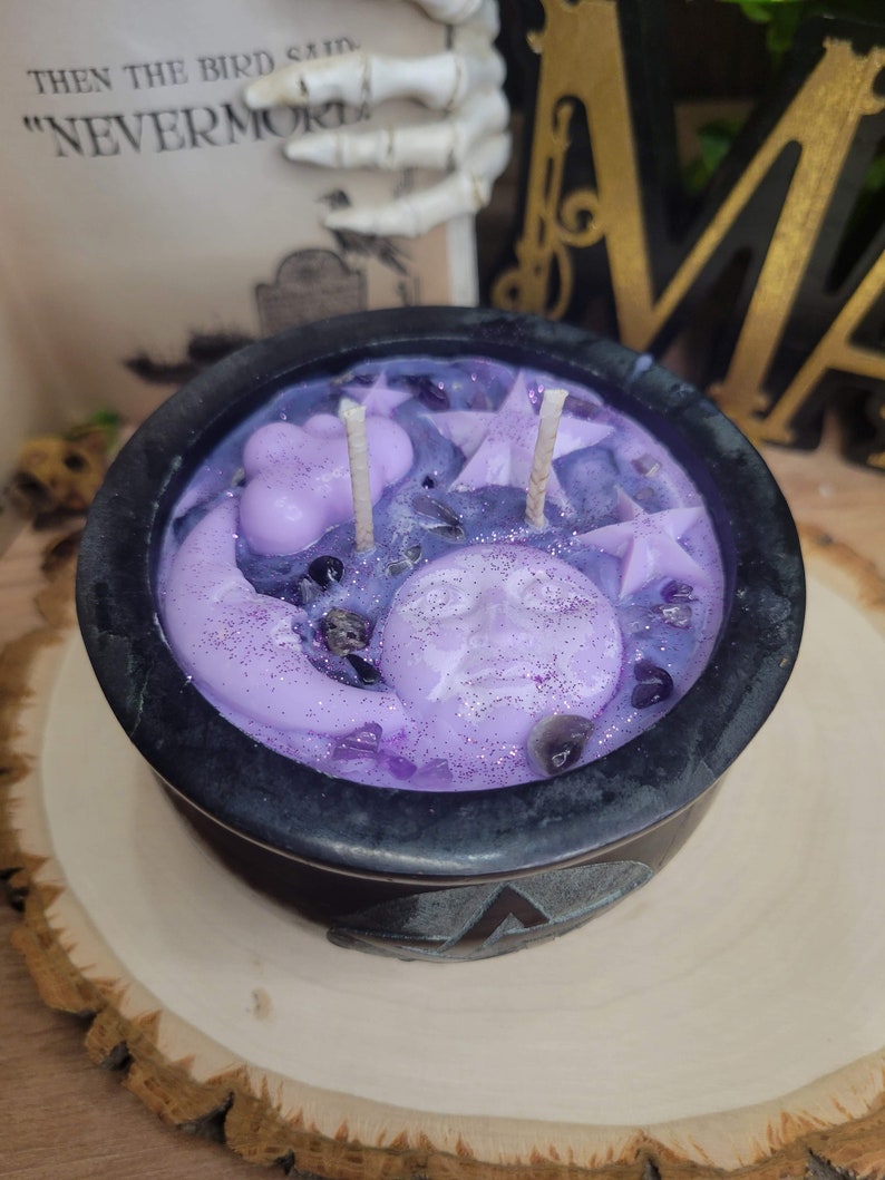 May include: A black cauldron-shaped candle with a lavender-colored wax. The candle features a moon, stars, and cloud-shaped wax accents. The candle sits on a wooden surface. The text "THEN THE BIRD SAID "NEVERMORE" is visible in the background.