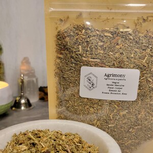 May include: A close-up of a bag of dried Agrimony herb. The bag is labeled with the name of the herb, its magical properties, and its gender, planet, element, and powers. The herb is green and brown and looks like it is ready to be used for herbalism or witchcraft.