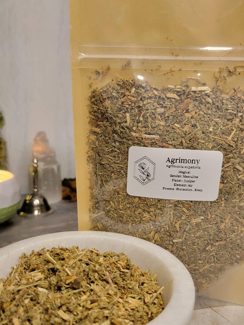 May include: A close-up of a bag of dried Agrimony herb. The bag is labeled with the name of the herb, its magical properties, and its gender, planet, element, and powers. The herb is green and brown and looks like it is ready to be used for herbalism or witchcraft.