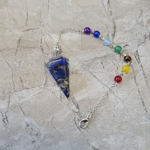 May include: A silver chain with a blue and white crystal pendulum and seven chakra beads. The beads are purple, blue, light blue, green, brown, yellow, and red.