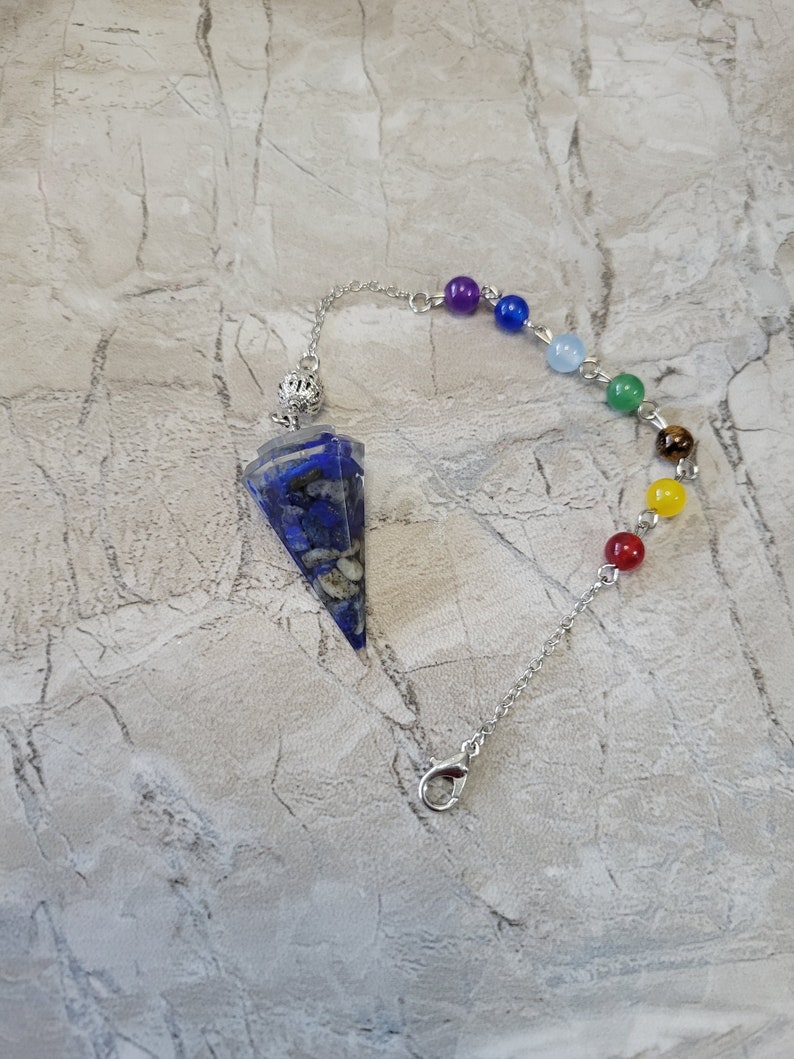 May include: A silver chain with a blue and white crystal pendulum and seven chakra beads. The beads are purple, blue, light blue, green, brown, yellow, and red.