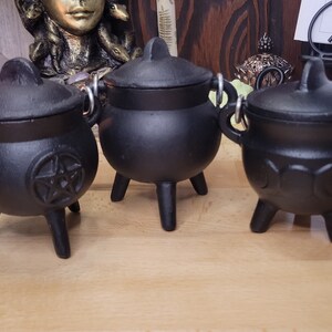 May include: Three small, black cast iron cauldrons with lids and handles. The cauldrons have three legs and are designed with different symbols, including a pentagram and crescent moons. The cauldrons are likely used for decorative or ritual purposes.