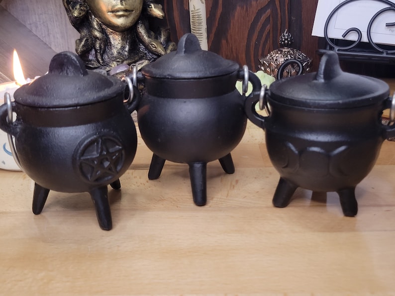 May include: Three small, black cast iron cauldrons with lids and handles. The cauldrons have three legs and are designed with different symbols, including a pentagram and crescent moons. The cauldrons are likely used for decorative or ritual purposes.