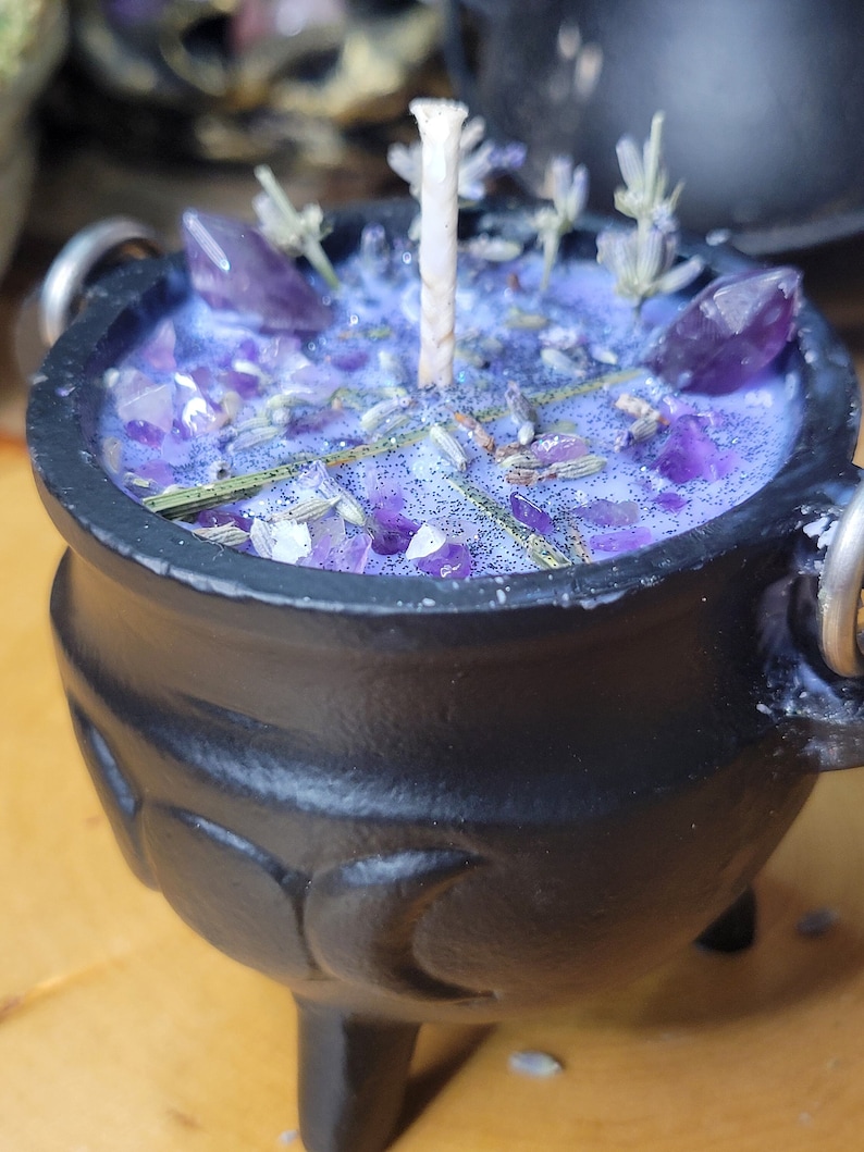 May include: A black cauldron-shaped candle with a purple wax interior. The candle is decorated with amethyst crystals, lavender sprigs, and glitter. A white wick rises from the center. The cauldron has three legs and two metal handles.