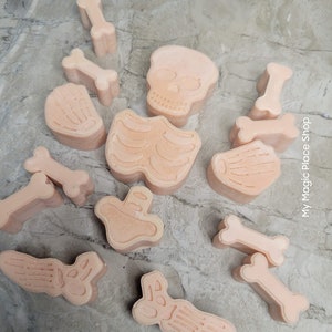 May include: A collection of light peach-colored, bone-shaped soaps and a skull-shaped soap are arranged on a gray surface. The soaps include rib cages, foot bones, and various other bone shapes, perfect for Halloween or novelty gifts.