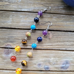 May include: A pair of long, silver chain earrings with colorful gemstone beads. The beads are arranged in a rainbow pattern, starting with red at the bottom and ending with purple at the top.