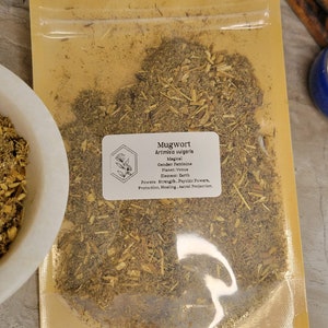 May include: A close-up of a resealable bag filled with dried mugwort herb. The bag is labeled with the name "Mugwort" and the botanical name "Artemisia vulgaris". The label also includes information about the herb's magical properties, including its gender, planet, element, and powers.