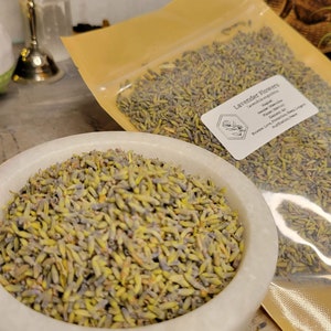 May include: A white ceramic bowl filled with dried lavender flowers. The flowers are a light purple and green colour. The bowl is sitting on a wooden surface. A clear plastic bag with a label that reads "Lavender Flowers" is in the background.